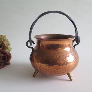 Pode incluir: Um caldeirão de cobre martelado de formato redondo com uma abertura larga, apresentando uma alça giratória de metal escuro fixada a dois anéis laterais. O recipiente é sustentado por três pequenos pés pontiagudos de cor latão.