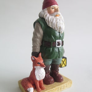 May include: A figurine of a gnome wearing a red hat and green jacket, holding a lantern and standing next to a red fox. The base of the figurine is brown and has the text "WWF 1997" on it.