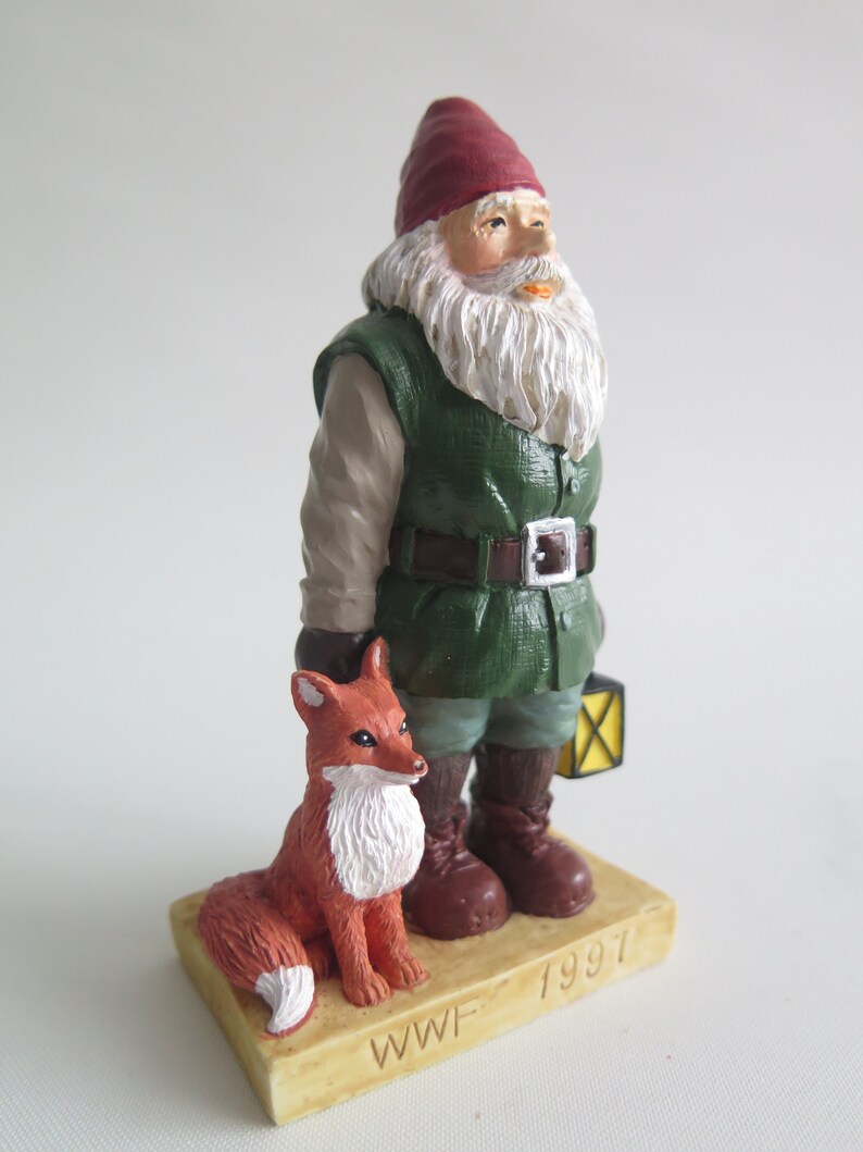May include: A figurine of a gnome wearing a red hat and green jacket, holding a lantern and standing next to a red fox. The base of the figurine is brown and has the text "WWF 1997" on it.
