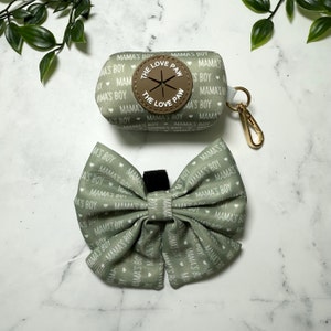 May include: A green fabric dog poop bag holder with a brown leather patch that says "The Love Paw" and a matching green fabric bow tie with white hearts and the text "Mama's Boy" repeated.