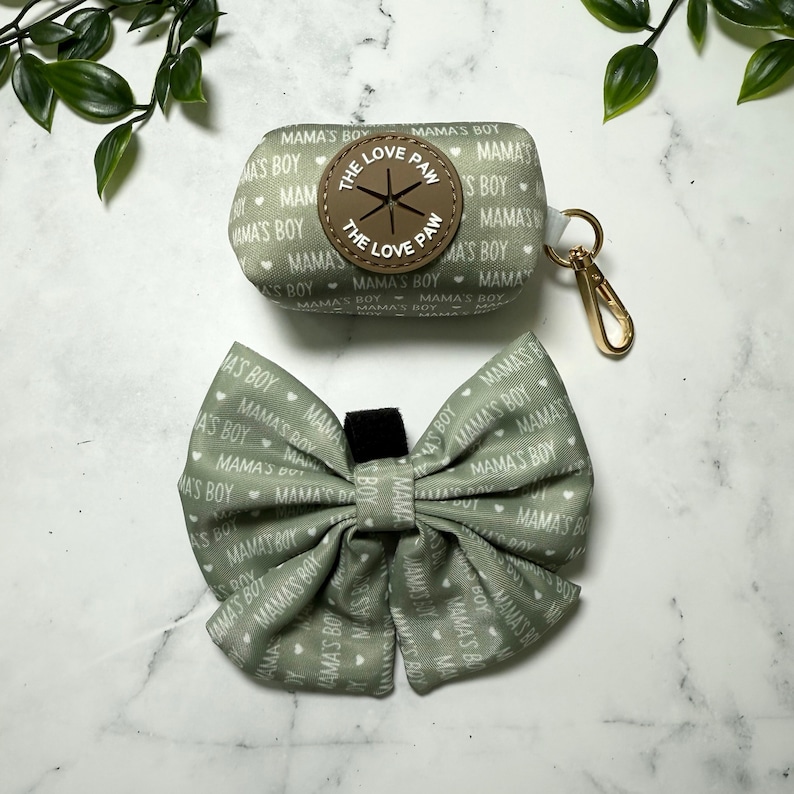 May include: A green fabric dog poop bag holder with a brown leather patch that says "The Love Paw" and a matching green fabric bow tie with white hearts and the text "Mama's Boy" repeated.