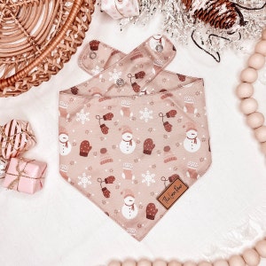 Può includere: Un bandana rosa con un motivo invernale con pupazzi di neve, guanti e fiocchi di neve. Il bandana ha un'etichetta in pelle marrone con il testo "The Love Paw".