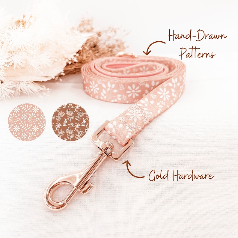 Op de afbeelding: Een roze hondenriem met een wit bloemenpatroon en gouden hardware. De riem is opgerold en de hardware is zichtbaar. De tekst "Handgetekend patronen" en "Gouden hardware" staat op de afbeelding.