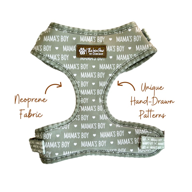 May include: A gray dog harness with a repeating pattern of white hearts and the text "Mama's Boy". The harness is made of neoprene fabric and features a unique hand-drawn pattern. The harness has a label that reads "The Love Paws Pet Lover Shop".