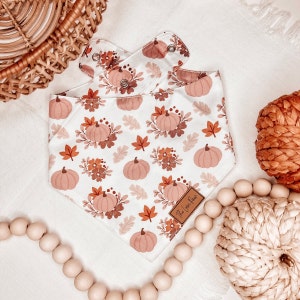 Puede incluir: Un pañuelo blanco con un estampado de otoño con hojas naranjas y marrones y calabazas. El pañuelo tiene una etiqueta de cuero marrón con el texto "The Love Nest" en él.