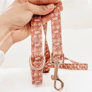 Op de afbeelding: Een bruine hondenriem met een wit bloemenpatroon en een gouden metalen gesp.