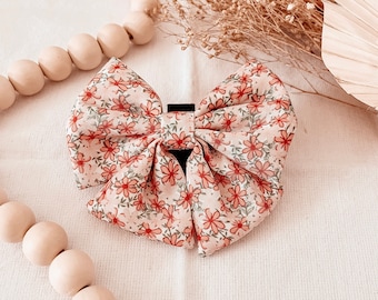 May include: A floral patterned hair bow with a black clip. The bow features a light pink background with red and green floral details. The bow is a large, decorative accessory.