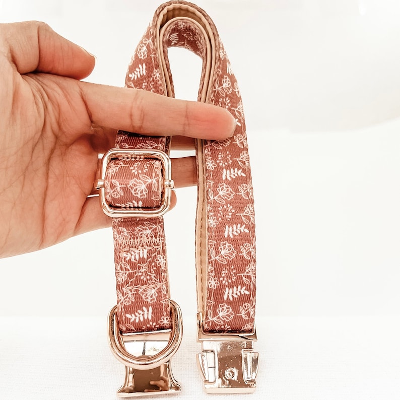 Puede incluir: Un collar para perro de color rosa y blanco con estampado floral y herrajes dorados. El collar tiene un cierre de hebilla y un anillo en D para sujetar una correa.