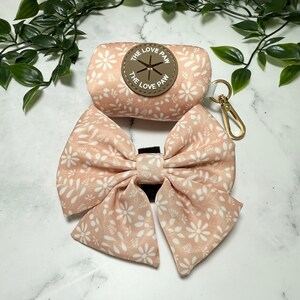 May include: A pink and white floral print dog poop bag dispenser with a matching bow. The dispenser has a brown leather patch with the text "The Love Paw" on it.