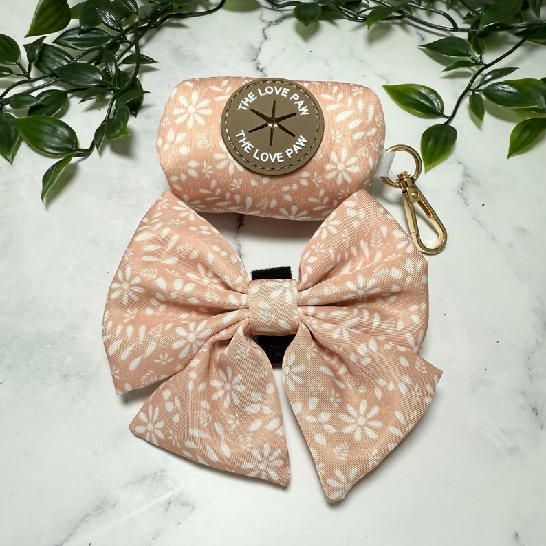 May include: A pink and white floral print dog poop bag dispenser with a matching bow. The dispenser has a brown leather patch with the text "The Love Paw" on it.