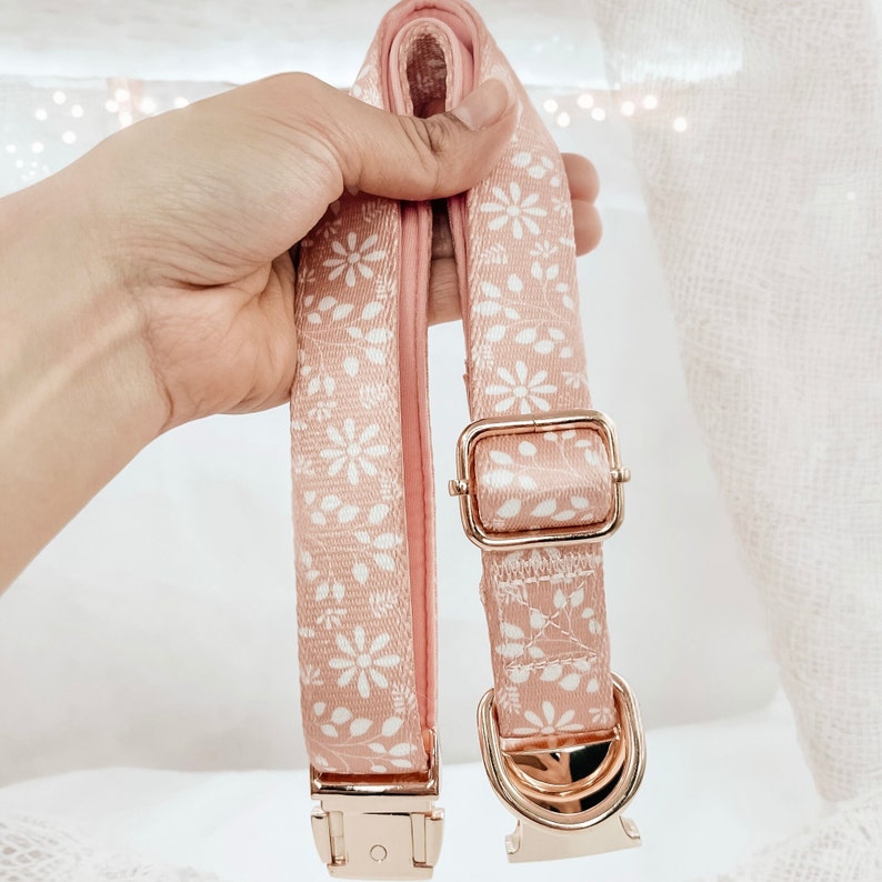 Puede incluir: Un collar para perro rosa con un patr&oacute;n floral blanco y herrajes de metal dorado.