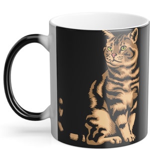 Puede incluir: Una taza que cambia de color con una ilustración detallada de un gato atigrado. El gato tiene rayas marrones y negras, ojos verdes y una mirada enfocada. El asa de la taza es negra y el interior es blanco.