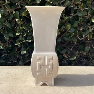 May include: A tall, off-white ceramic vase with a flared top and a rectangular base. The vase features a geometric design on the front and is set against a backdrop of green foliage.