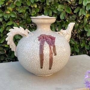 May include: A white ceramic teapot with a dragon handle and a crackled glaze. The teapot has a red drip glaze on the side.