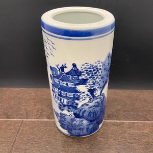 May include: A cylindrical white and blue ceramic vase. The vase features a detailed blue landscape design with buildings and trees. The top and base have blue bands. The vase is standing on a wooden surface.