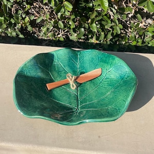 May include: A green ceramic serving dish shaped like a leaf with a wooden spreader. The dish has a textured surface that resembles a leaf.
