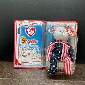 May include: A Ty Beanie Baby bear named Spangle, packaged in a clear plastic case. The bear is white with a red and white striped bow and red, white, and blue star-spangled clothing. The packaging includes text and a McDonald's logo.