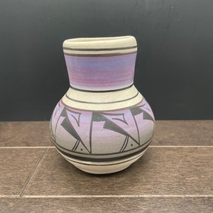 May include: A small, white ceramic vase with a purple and black geometric design. The vase has a rounded body and a narrow neck.