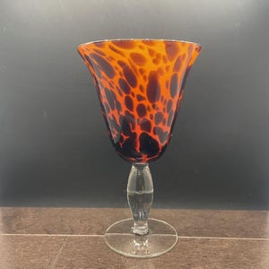 May include: A tall, elegant wine glass with a flared rim. The glass features a striking pattern of orange and black spots, resembling tortoiseshell. The stem and base are clear, adding to the glass's sophisticated design.
