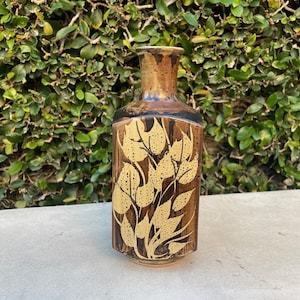 May include: A brown ceramic vase with a rectangular body and a narrow neck. The vase features a gold leaf pattern on a dark brown background. The rim and neck are a lighter brown color. The vase is approximately 10 inches tall.
