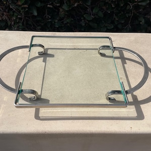 May include: A silver tray with two handles and a glass top. The tray is square and has a simple design.