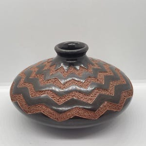 May include: A dark brown ceramic vase with a textured, zigzag pattern in a lighter brown color. The vase has a rounded body and a narrow, dark brown opening. The design is carved into the surface.