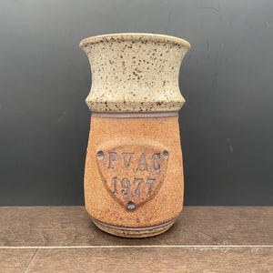 May include: A brown and white ceramic vase with a speckled glaze. The vase has a rounded base and a flared top. It has a brown shield-shaped design with the text "PVAG 1977" on the front.