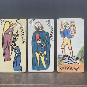 Puede incluir: Tres baldosas cerámicas rectangulares con ilustraciones religiosas. Una representa un ángel y el texto "Ste Gabrielle", otra muestra a un hombre con un libro y el texto "St Matthieu", y la última muestra a un hombre que lleva a un niño con el texto "St Christophe".