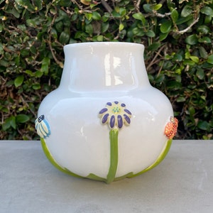 May include: White ceramic vase with a round body and a wide mouth. The vase is decorated with three green stems with flowers and butterflies. The flowers are purple, yellow, and white. The butterflies are blue, red, and yellow.