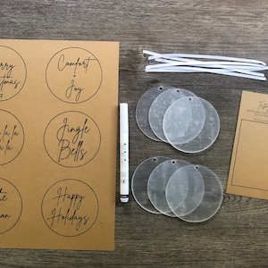 May include: A set of six blank acrylic ornaments, a sheet of paper with six Christmas-themed designs, a white marker, and a sheet of paper with a "Tasty Party" heading and a floral design. The ornaments are ready to be personalized with the designs.