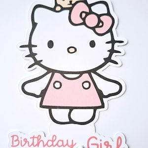 May include: A white and pink Hello Kitty cake topper with a small bear on top. The topper has a pink bow and dress, black outlines, and the words "Birthday Girl" in pink script.