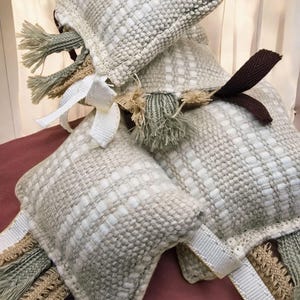 May include: A collection of square, woven decorative pillows in shades of beige and white. Each pillow features a textured pattern and is adorned with tassels, ribbons, and bows in complementary colors. The pillows are arranged in a casual, layered composition.