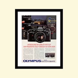 Peut inclure: Un appareil photo Olympus OM10FC Quartz noir avec un objectif argenté et un flash argenté. L'appareil photo est doté d'une sangle noire et argentée et est posé sur un fond blanc. Le texte "OLYMPUS OM10FC QUARTZ" est imprimé en blanc sur l'appareil photo.