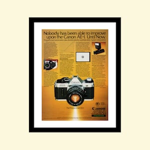 Peut inclure: Un appareil photo Canon AE-1 Programmed noir avec un objectif argenté. L'appareil photo est sur un fond blanc avec du texte qui dit "Personne n'a pu améliorer le Canon AE-1. Jusqu'à maintenant." et "Le Programmed AE-1".