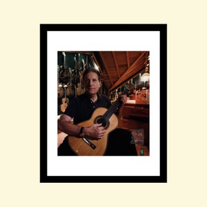 Puede incluir: Un hombre con una camisa negra toca una guitarra clásica en una habitación llena de otras guitarras. La habitación tiene un techo y paredes de madera.