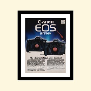 May include: A vintage advertisement for Canon EOS cameras, featuring two black cameras with red lenses. The text reads "Canon EOS System" and "More than autofocus. More than ever."