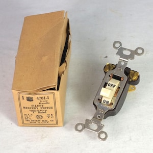 May include: An antique electrical mercury switch with an ivory handle, labeled "OFF" and "ON". The switch is mounted on a metal plate with screw holes. A cardboard box with product information is next to the switch.