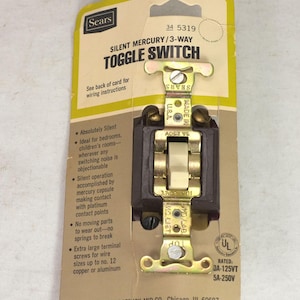 Könnte beinhalten: Vintage Sears Silent Mercury 3-Wege-Kippschalter auf Originalverpackung. Der Schalter hat ein braunes Gehäuse, einen cremefarbenen Kippschalter und goldfarbene Metallkomponenten. Die Verpackung ist gelb und braun.