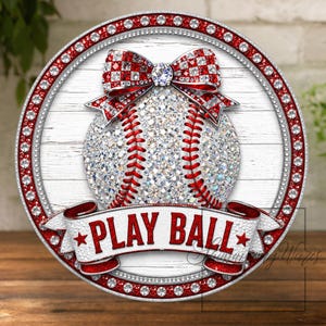 May include: A round baseball-themed decorative item with a sparkling baseball design, a red bow, and the words "PLAY BALL" on a banner. The border is adorned with red and clear rhinestones, set against a white wood-grain background.