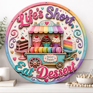 Può includere: Un cartello da parete circolare con una colorata illustrazione di un carrello dei dolci con cupcake, macaron e fette di torta. Il testo recita Life's Short Eat Dessert e Sweet Treats. Il design include colori pastello, cuori e ghirigori.