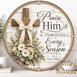 May include: Round wooden wall art with a rustic cross, cream-colored bow, and floral arrangement. The text reads "Praise Him Through Every Season" with the Bible verse Psalm 146:2. The artwork has a distressed white wood background.