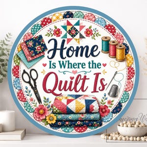 May include: A circular sign with a blue border, featuring the text "Home Is Where the Quilt Is." The design includes quilting motifs such as thread spools, scissors, and stacked quilts. The sign incorporates floral and patchwork patterns.
