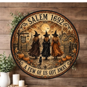May include: A round wall sign featuring a scene of three witches in pointed hats walking toward a village under a full moon. The text reads SALEM 1692 at the top and A FEW OF US GOT AWAY at the bottom. The image includes pumpkins and a black cat.