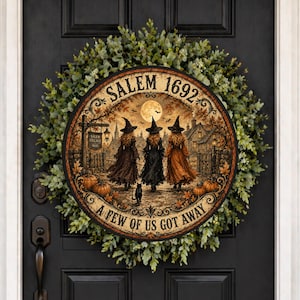 May include: A round wooden sign featuring a scene of three witches in pointed hats walking toward a village, with a full moon, pumpkins, and the text SALEM 1692 and A FEW OF US GOT AWAY, mounted on a lush green wreath on a black door.