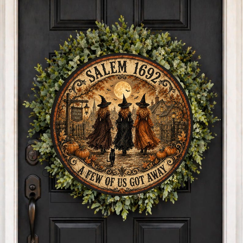 May include: A round wooden sign featuring a scene of three witches in pointed hats walking toward a village, with a full moon, pumpkins, and the text SALEM 1692 and A FEW OF US GOT AWAY, mounted on a lush green wreath on a black door.