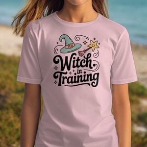 May include: A light pink t-shirt with the words "Witch in Training" in a whimsical font. The design includes a witch hat, a star-shaped wand, and decorative swirls. The shirt is a short-sleeved crew neck.