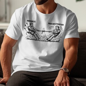 May include: White t-shirt featuring a black and white illustration of a tug-of-war. The design shows two figures, labelled "Progress" and "Procrastination", pulling on a rope, with graphic details.