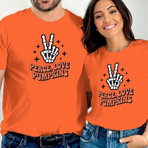 Könnte beinhalten: Leuchtend orangefarbene T-Shirts mit dem Text "Peace, Love, Pumpkins" und einer Skelett-Hand, die das Friedenszeichen zeigt. Das Design enthält Sterne. Die Shirts sind leuchtend orange.
