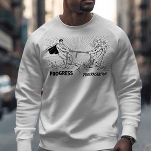 May include: White sweatshirt with a black and white illustration of two figures in a tug-of-war, with the words "PROGRESS" and "PROCRASTINATION". The design shows a battle between progress and procrastination.