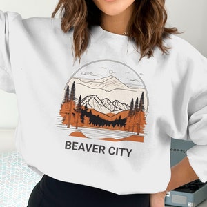 May include: White sweatshirt featuring a graphic of a mountain range with trees and a lake. The text "BEAVER CITY" is printed below the graphic.